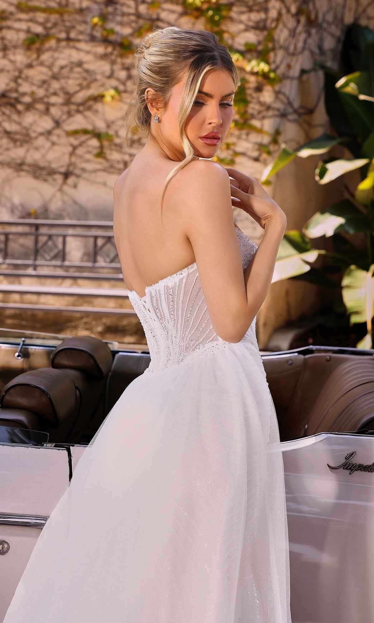 A woman in the Ladivine CC492W long formal dress, featuring a strapless white design with crystal beaded embellishments, stands beside a vintage white car and looks over her shoulder.